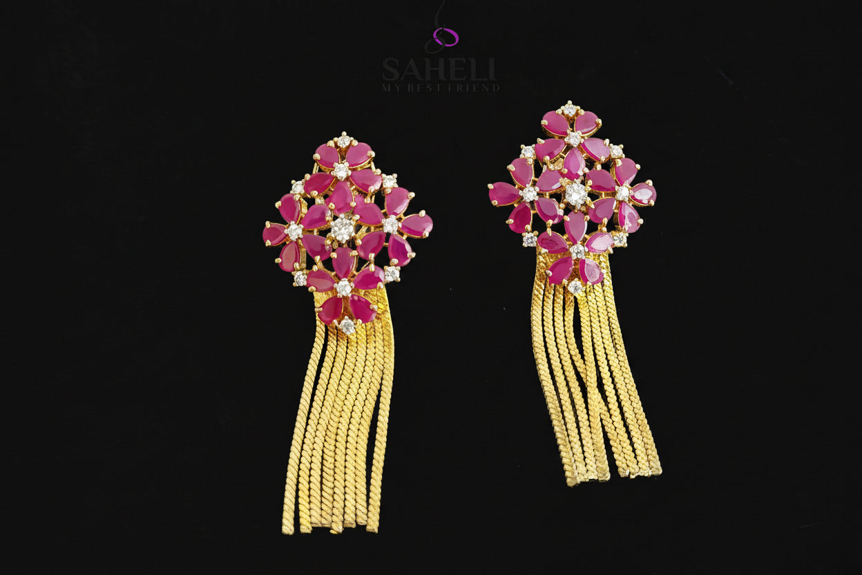 Pair of earrings with pink gemstones and yellow tassels on a dark background, branded 'Saheli'.