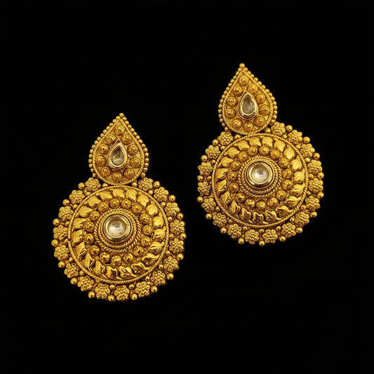 A pair of ornate gold-toned traditional Indian jhumka earrings with intricate filigree work and clear stone accents, displayed on a dark reflective surface with the SAHELI brand logo at the top.