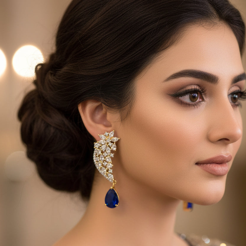 Gold and diamond earrings with blue teardrop pendants on a gray stand, branded 'SAHELI'.