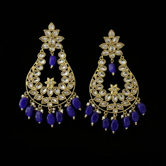 A pair of gold-plated Indian chandelier earrings with clear stone pavé settings and dangling royal blue beads, displayed on a dark reflective surface with the Saheli My Best Friend logo in the top left corner.