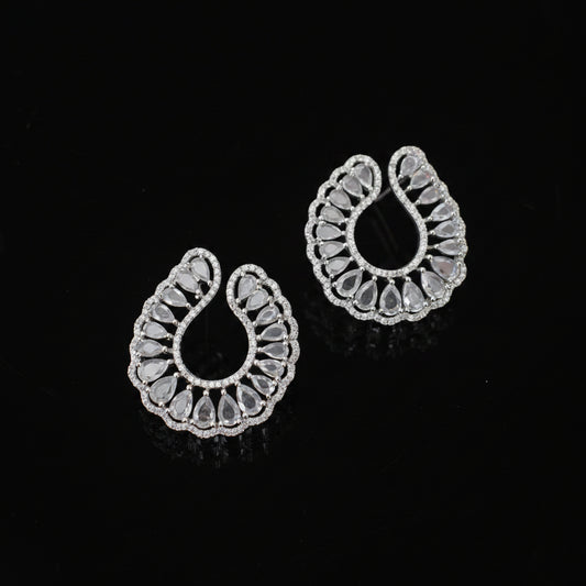 A pair of silver-toned teardrop-shaped earrings with intricate filigree cutouts and clear stones, displayed on a dark reflective surface.