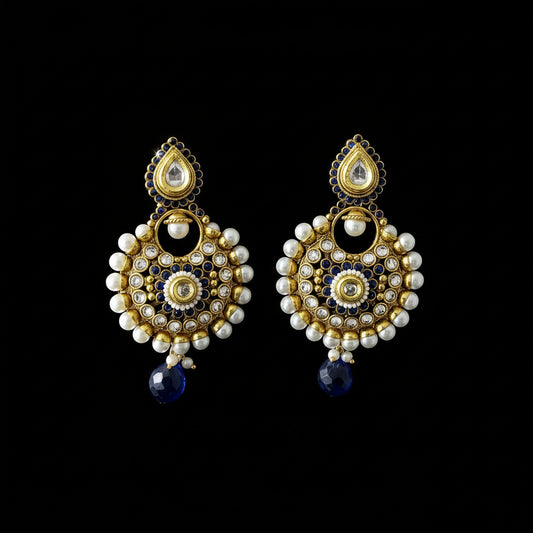 A pair of ornate Indian jhumka earrings with gold-plated metalwork, white pearl beads, blue enamel details, and dark blue teardrop dangling charms, displayed on a dark gray reflective surface with the 'SAHELI MY BEST FRIEND' logo at the top.