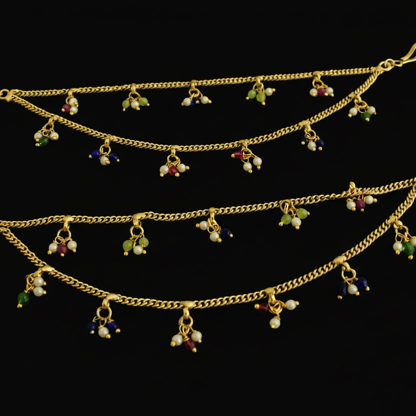 Two gold anklets with colorful charms on a dark background