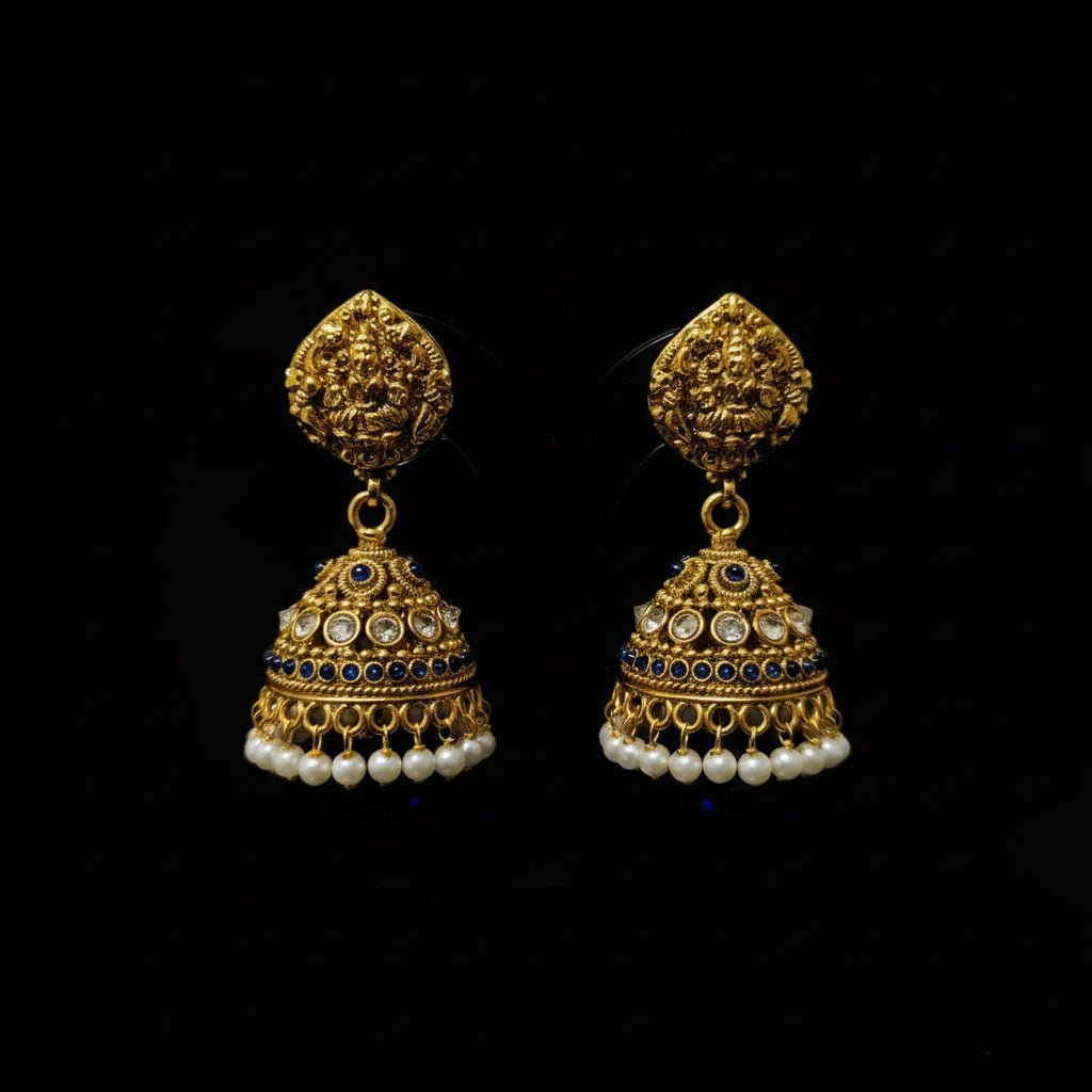 A pair of ornate gold-toned jhumka earrings displayed on a clear stand, with a small 'Saheli Jewellery' tag attached. The earrings feature intricate gold filigree, dark blue stone accents, and rows of white faux pearls at the bottom, set against a blurred desk background with a laptop keyboard.