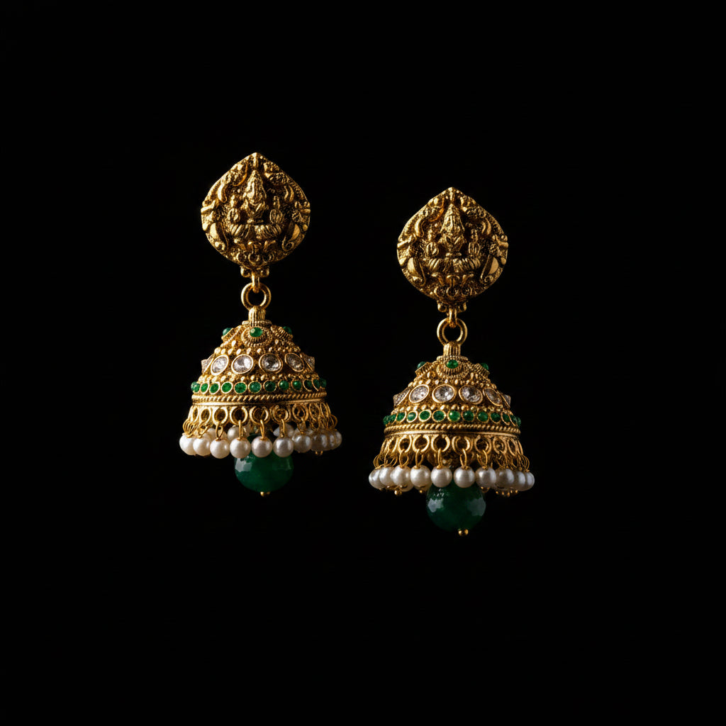 A pair of ornate gold-toned Indian jhumka earrings with green stone accents, white pearl bead drops, and a textured gold top, displayed on a black Y-shaped stand against a gray background. The brand logo 'SAHELI MY BEST FRIEND' is visible at the top center.