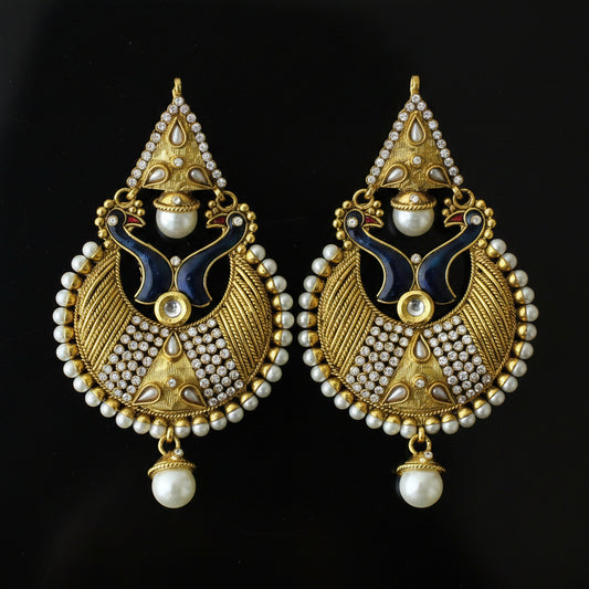 A pair of traditional Indian gold-plated earrings with intricate filigree, white faux pearls, and dark blue enamel details, displayed on a dark reflective surface. The Saheli My Best Friend logo is visible in the top left corner.