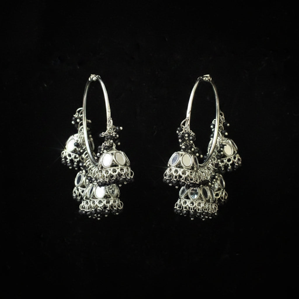 A pair of silver-toned traditional hoop earrings with dangling ornate jhumka-style charms and mirror-like stones, displayed on a black background.