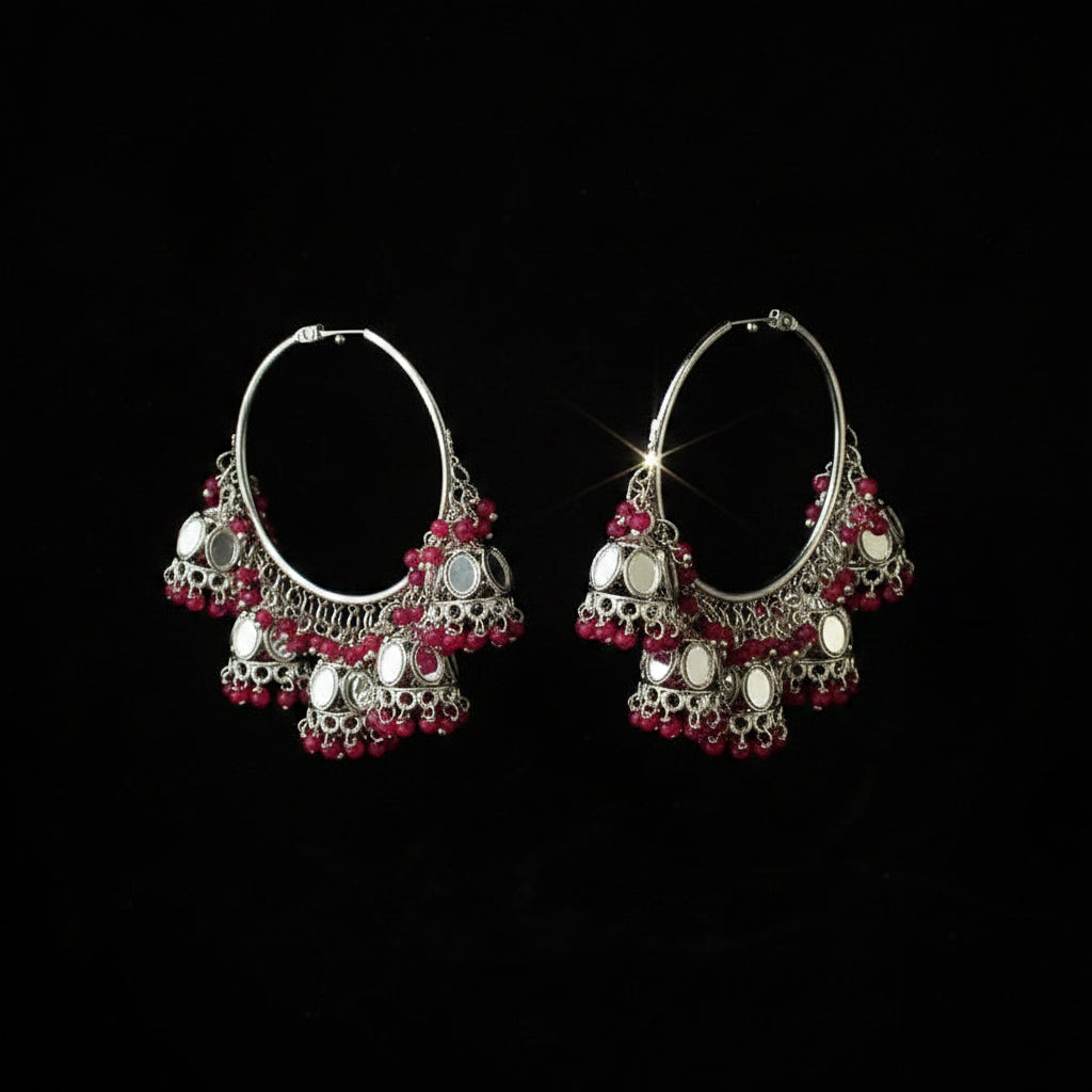 A pair of silver-toned hoop earrings with dangling filigree details, red bead accents, and mirrored circular elements, displayed against a solid black background.