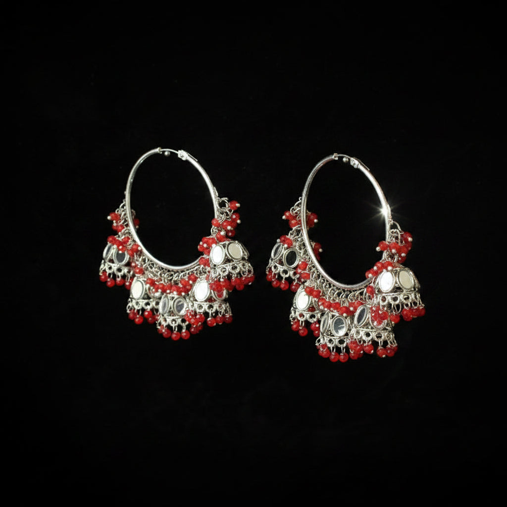 A pair of silver-toned hoop earrings decorated with intricate filigree, small red beads, and round silver mirror-like discs, displayed against a solid black background.