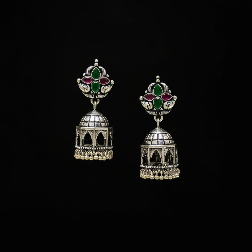 Silver jhumka earrings with green and red stones on a black stand against a gray background.