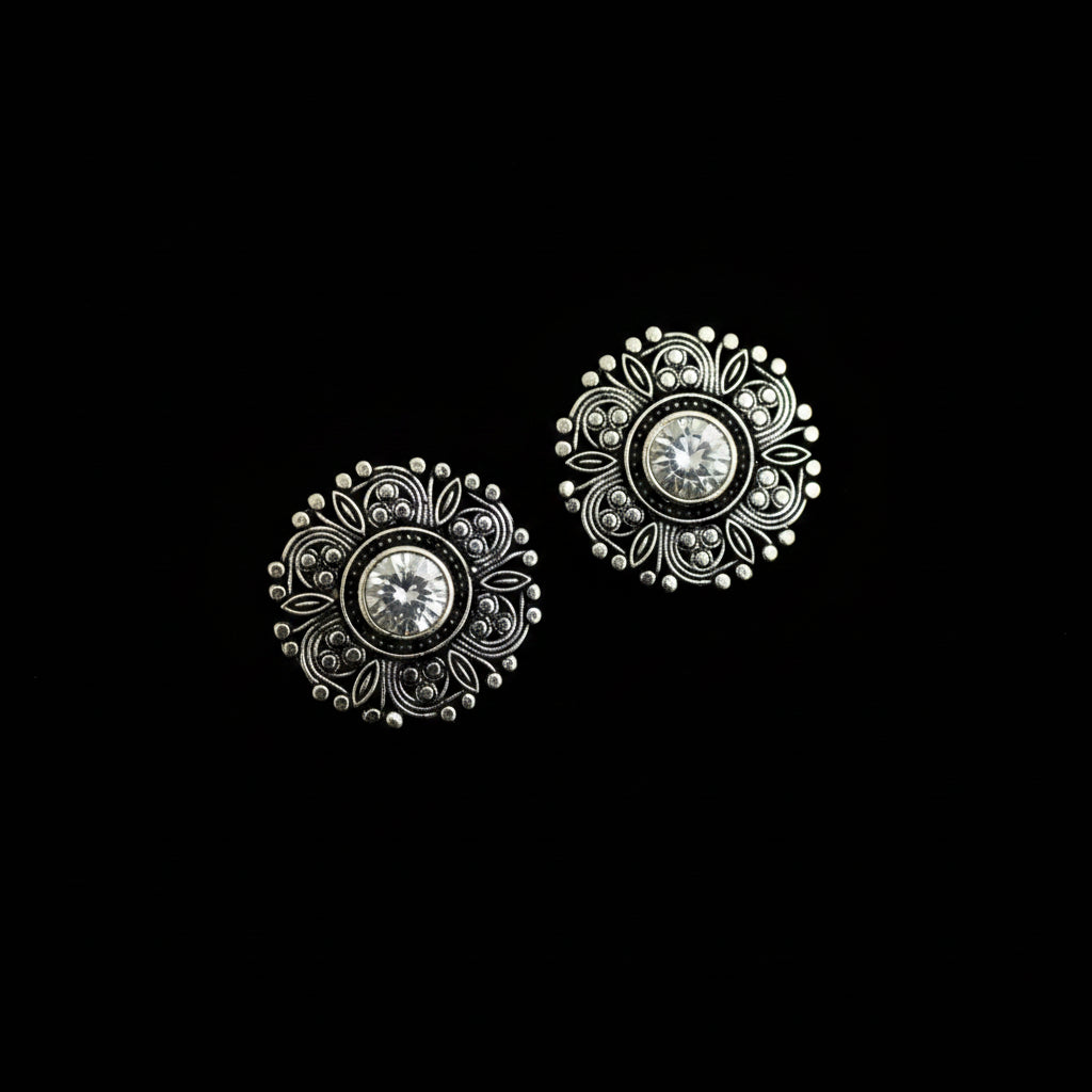 A pair of ornate silver-toned filigree stud earrings with clear round gemstones, displayed on a dark reflective surface.