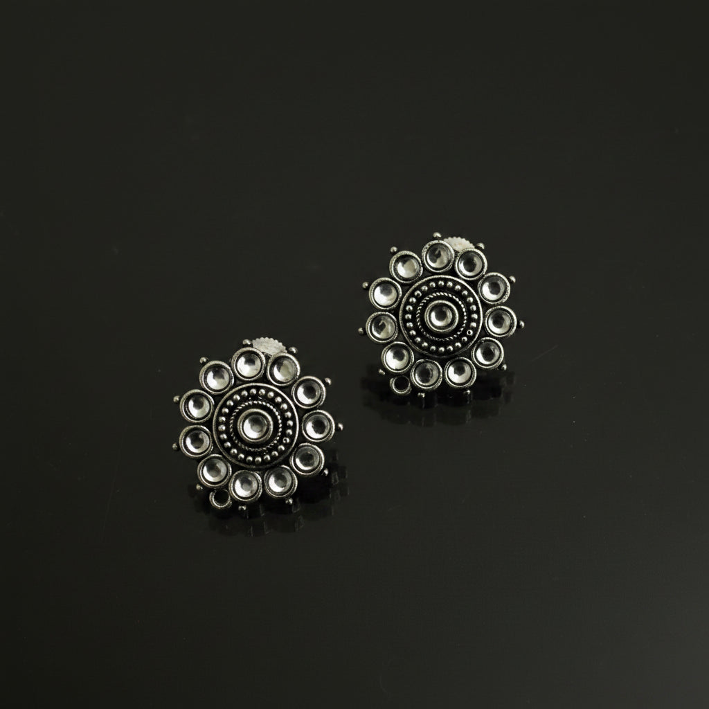 A pair of silver-toned Indian Kundan earrings with intricate circular floral designs and small clear stones, displayed on a dark reflective surface.