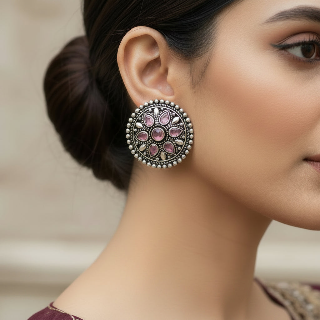 Two round silver-toned decorative jewelry pieces with pink teardrop and circular stones, displayed on a dark reflective surface.