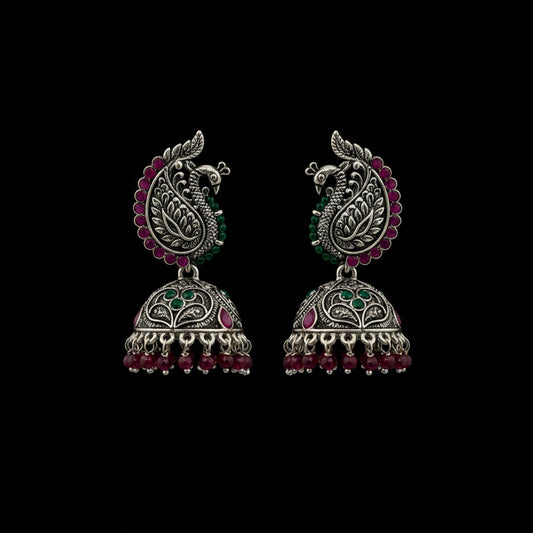 A pair of ornate silver-toned Indian jhumka earrings with peacock motifs, pink and green stone accents, and dangling bead chains, displayed on a black jewelry stand against a dark gray background.