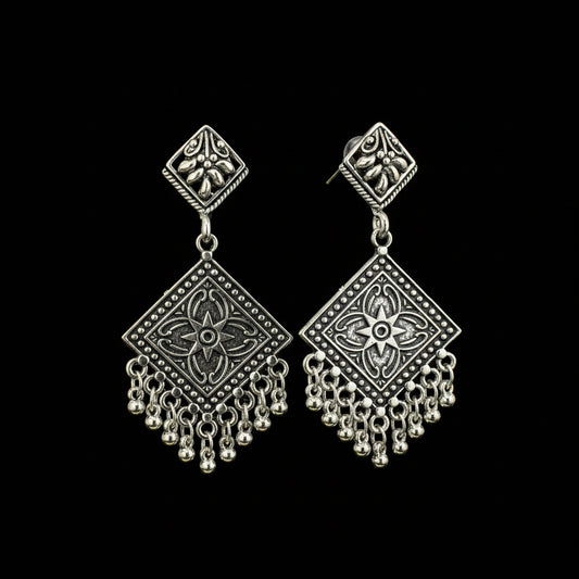 A pair of silver-toned geometric dangle earrings with ornate filigree and bead fringe, displayed on a black Y-shaped jewelry stand against a dark gray background.