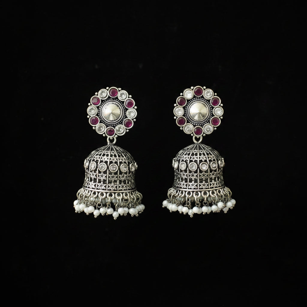 A pair of ornate silver-toned Indian jhumka earrings displayed on a black Y-shaped jewelry stand against a black background. The earrings feature circular floral studs with red stones and a central silver stone, hanging from bell-shaped dangles decorated with silver filigree and rows of small white pearl-like beads at the bottom.