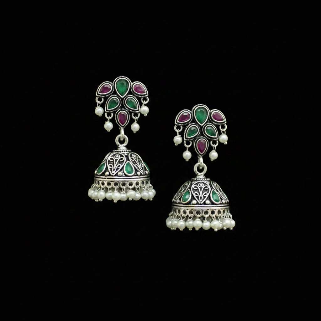 A pair of traditional Indian silver jhumka earrings with green and purple enamel stones and white pearl bead borders, displayed on a black Y-shaped jewelry stand.