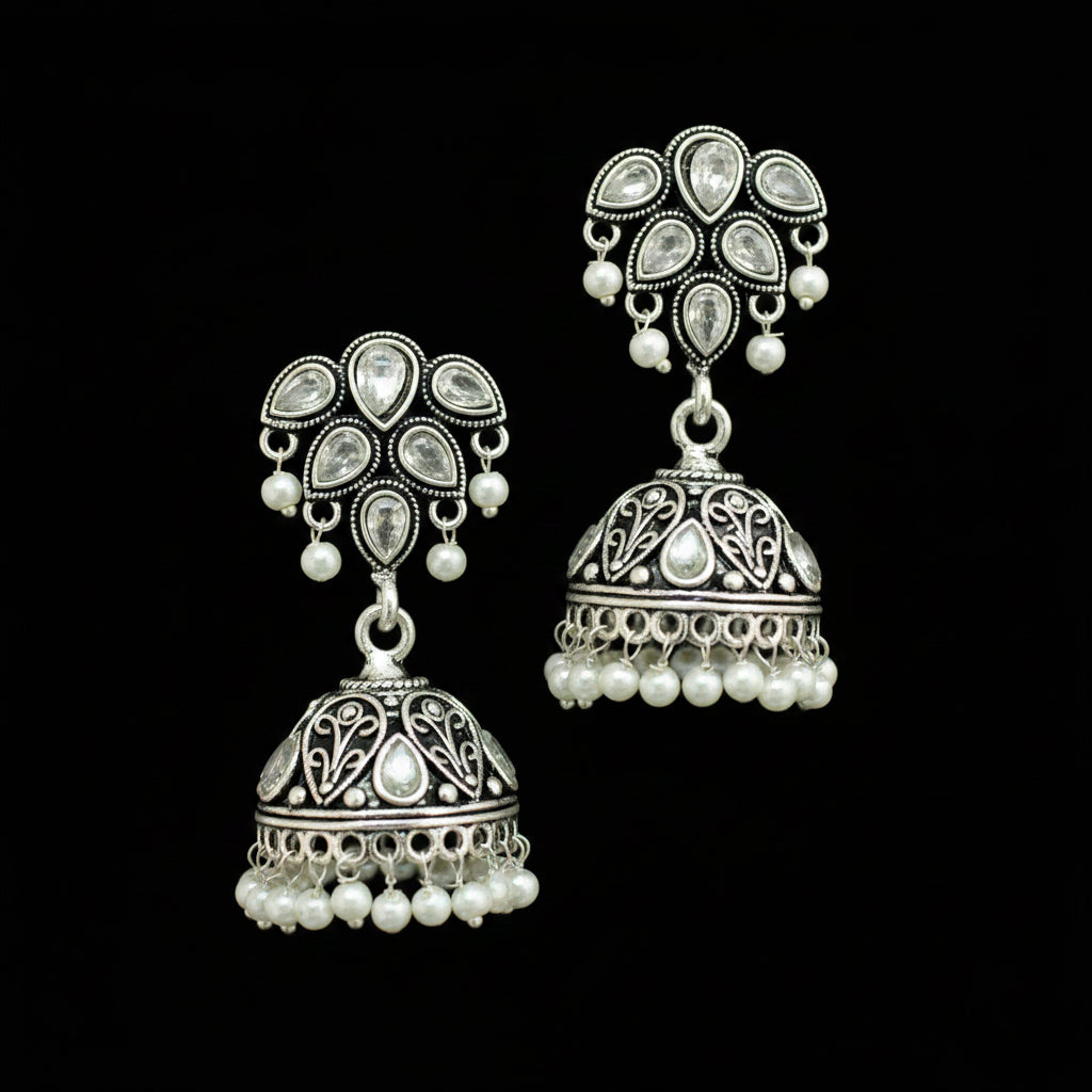 A pair of silver-toned Indian jhumka earrings with white pearl bead accents, displayed on a black jewelry stand against a gray background.