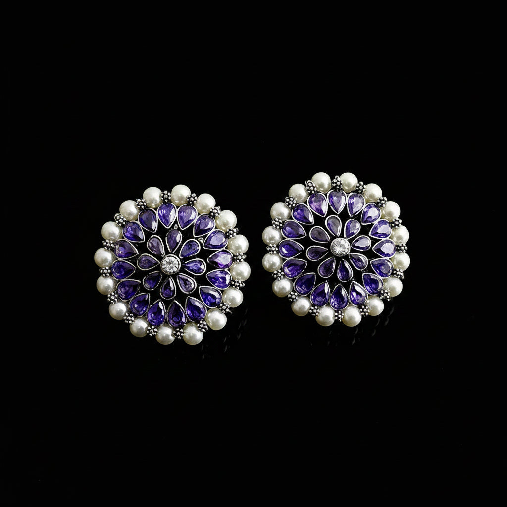 A pair of round floral stud earrings featuring purple amethyst teardrop stones, a central clear crystal, and a border of white faux pearls on silver-toned settings, displayed on a dark reflective surface with the SAHELI brand logo above.