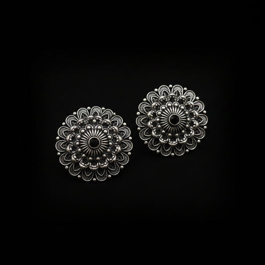 A pair of round, ornate silver-toned floral stud earrings with black oxidized detailing, displayed on a clean white background with the brand logo 'SAHELI MY BEST FRIEND' above them.