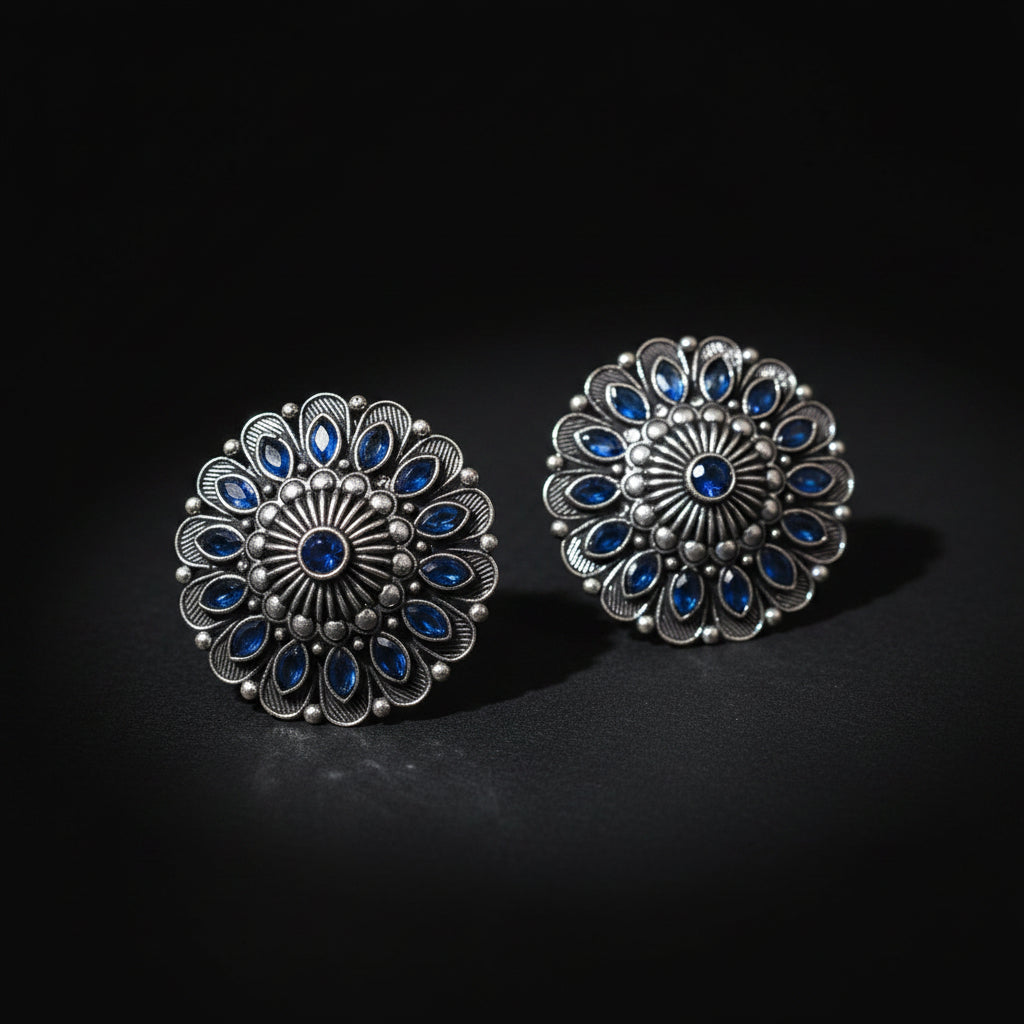 A pair of circular silver-toned brooches with intricate floral detailing and blue stone accents, displayed on a white background with the SAHELI MY BEST FRIEND logo above them.