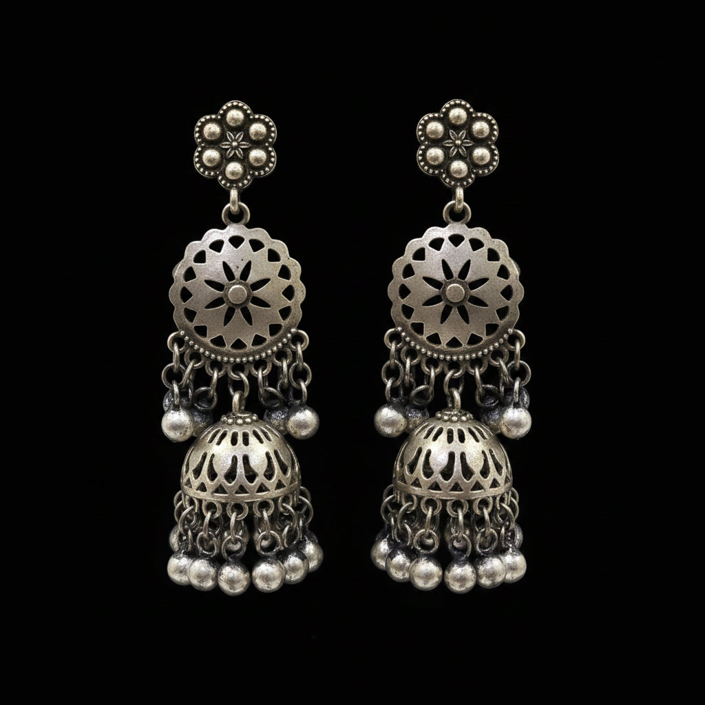 A pair of silver-toned Indian jhumka earrings displayed on a white stand. The earrings feature intricate filigree floral studs, circular patterns, and dangling bell-shaped jhumka charms with small silver beads at the bottom.