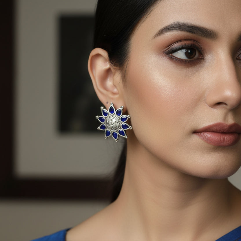 Pair of sun-shaped earrings with blue accents on a blue fabric background with 'Al-Rawnaq' branding.