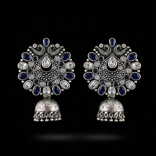 A pair of ornate silver-toned Indian jhumka earrings decorated with dark blue enamel and clear rhinestones, displayed on a black jewelry stand.