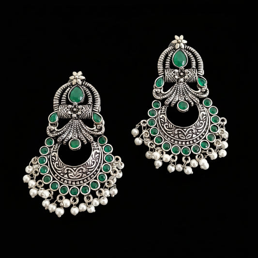 A pair of ornate silver-toned Indian jhumka earrings with green stones and white pearl-like bead drops, displayed on a dark reflective surface. The brand logo 'SAHELI BY SAHELI BRAND' is visible at the top center.