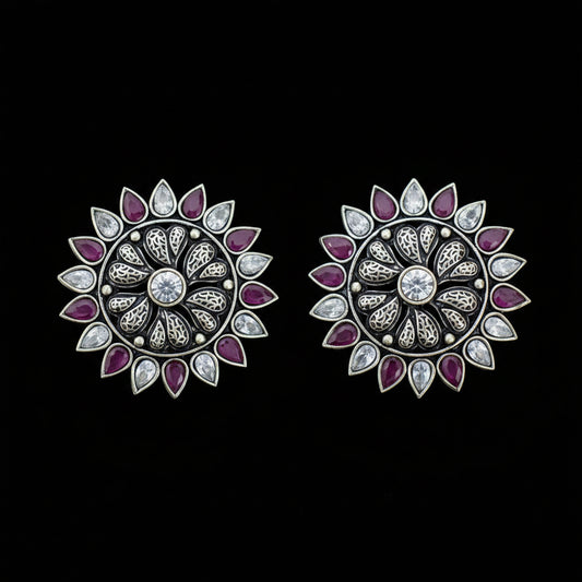 A pair of silver-toned floral stud earrings with purple gemstone petals and clear central stones, displayed on a dark gray reflective surface.