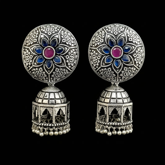 A pair of ornate silver-toned Indian jhumka earrings displayed on a gray Y-shaped jewelry stand. The earrings feature circular studs with intricate floral filigree, blue enamel accents, and a central pink stone, hanging from silver bell-shaped dangles with small pearl-like beads at the bottom.