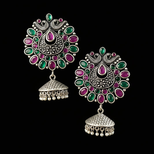 A pair of ornate Indian silver-toned jhumka earrings with green and pink stones, displayed on a gray stand against a neutral gray background with the brand name 'SAHELI MY BEST FRIEND' at the top.