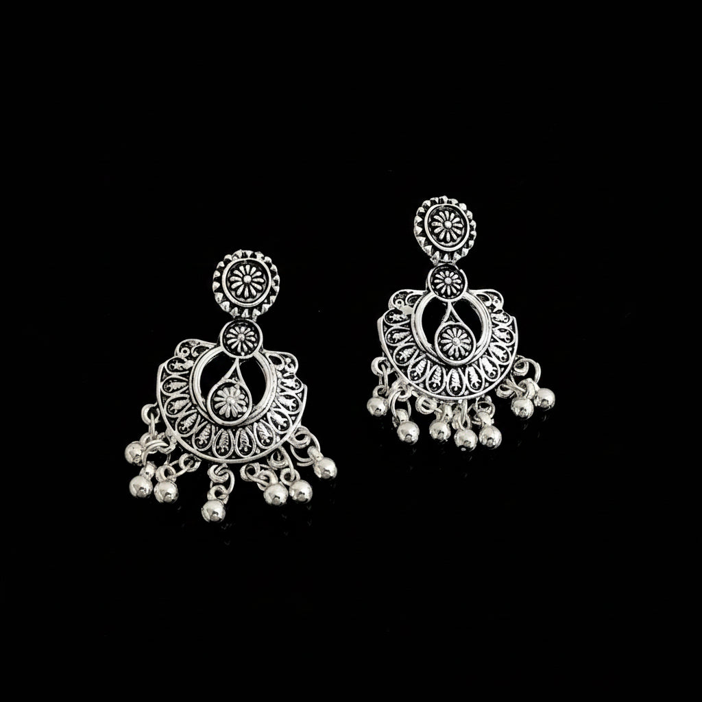 A pair of silver-toned Indian jhumka earrings with intricate filigree work and dangling bead chains, displayed on a dark reflective surface.