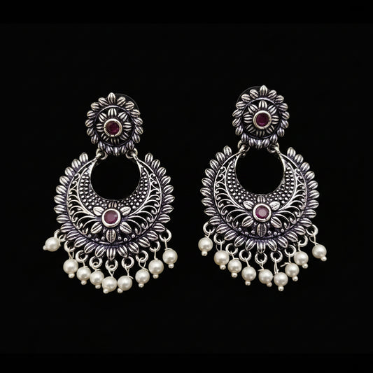 Pair of silver earrings with red stones and pearl drops on a gray background, branded 'Saheli'.
