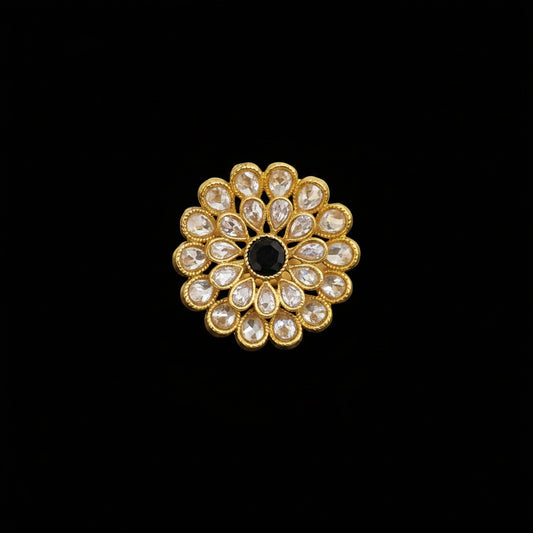 A gold flower-shaped ring with clear kundan stones and a central black stone, displayed on a reflective gray surface. The brand logo 'SAHELI MY BEST FRIEND' is visible in the top left corner.