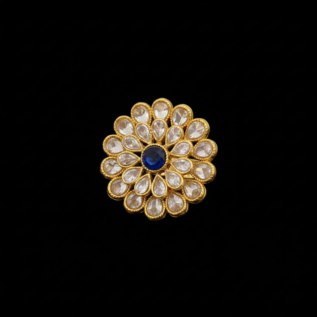 A gold-toned floral ring with clear teardrop-shaped stones and a central blue oval stone, displayed on a dark reflective surface with the SAHELI MY BEST FRIEND logo in the top left.