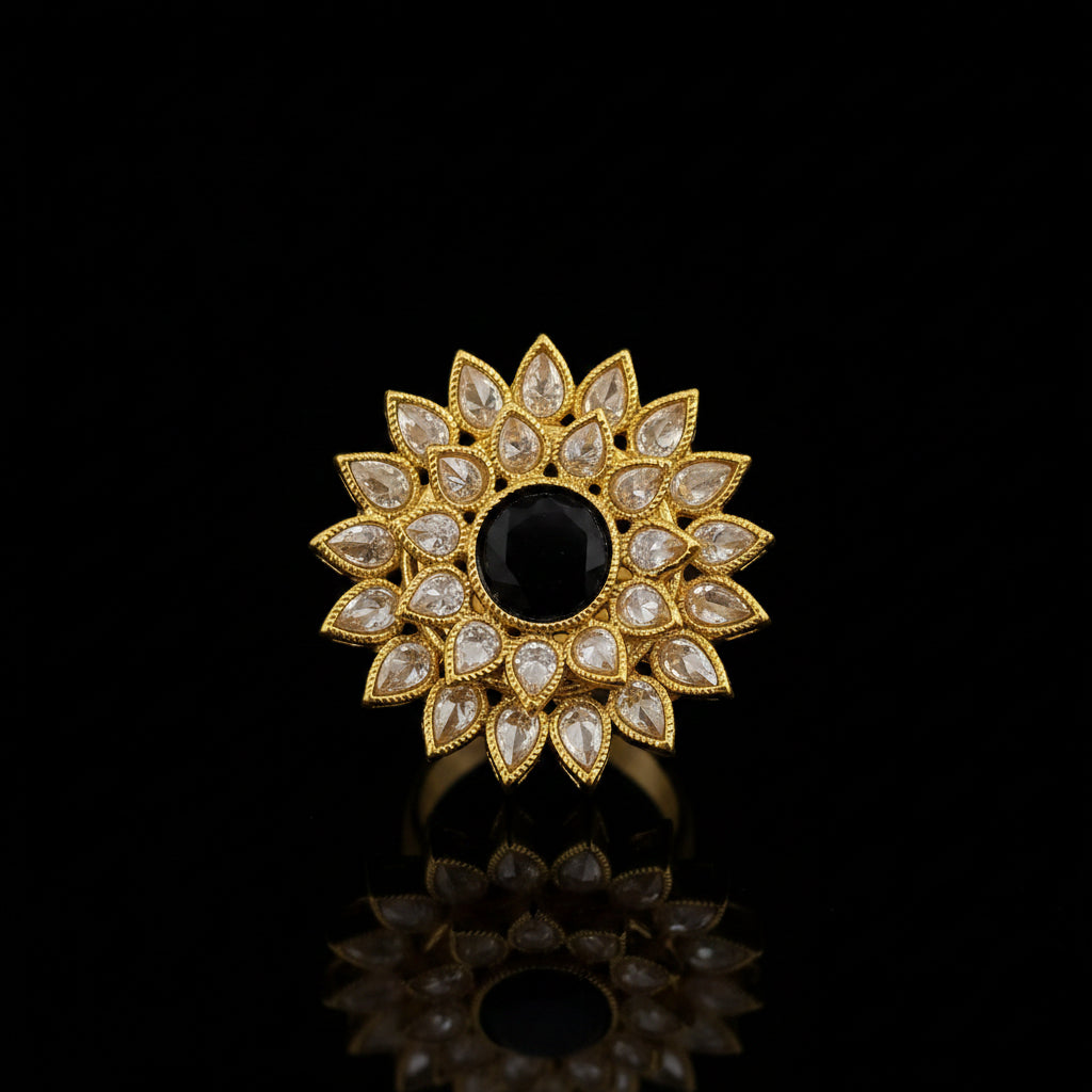 A gold-toned floral brooch featuring a central black oval stone surrounded by clear teardrop-shaped stones, displayed on a dark reflective surface with the SAHELI brand logo in the top left corner.
