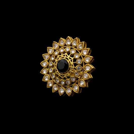 A gold-plated Kundan ring featuring a central black stone surrounded by clear stones in a floral mandala pattern, displayed on a reflective gray surface.