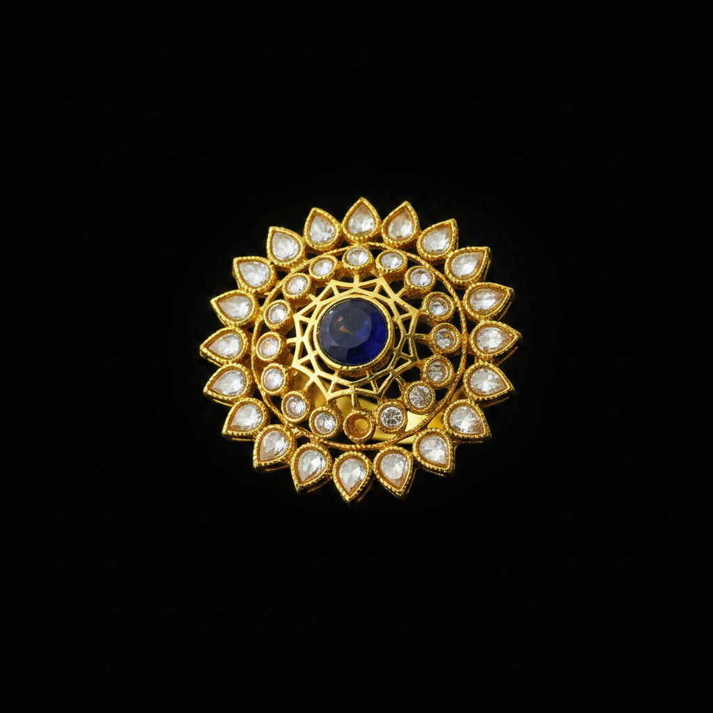 A gold-toned circular brooch or decorative pin featuring a central deep blue oval stone surrounded by clear teardrop-shaped stones, displayed on a dark gray background with the SAHELI MY BEST FRIEND logo in the top left.