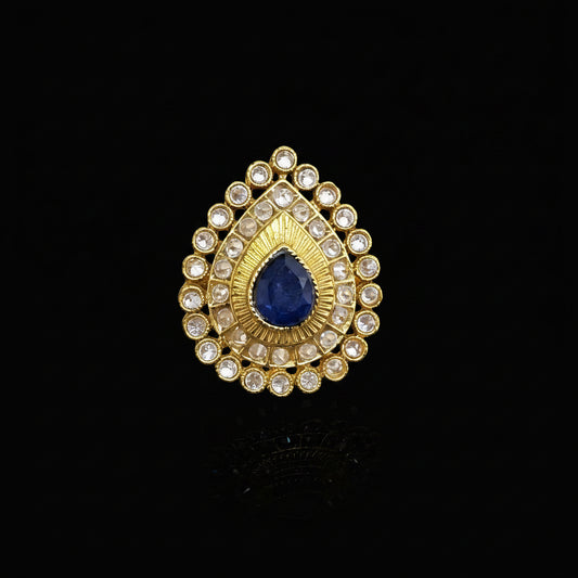 A gold teardrop-shaped ring with a central deep blue teardrop gemstone and surrounding clear rhinestones, displayed on a reflective gray surface. The brand logo 'SAHELI MY BEST FRIEND' is visible in the top left corner.
