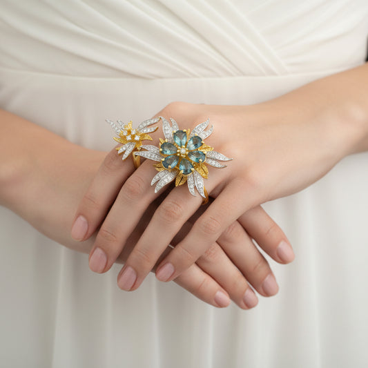 Gold and silver floral ring with gemstones on a gray background, featuring the brand 'Saheli'.