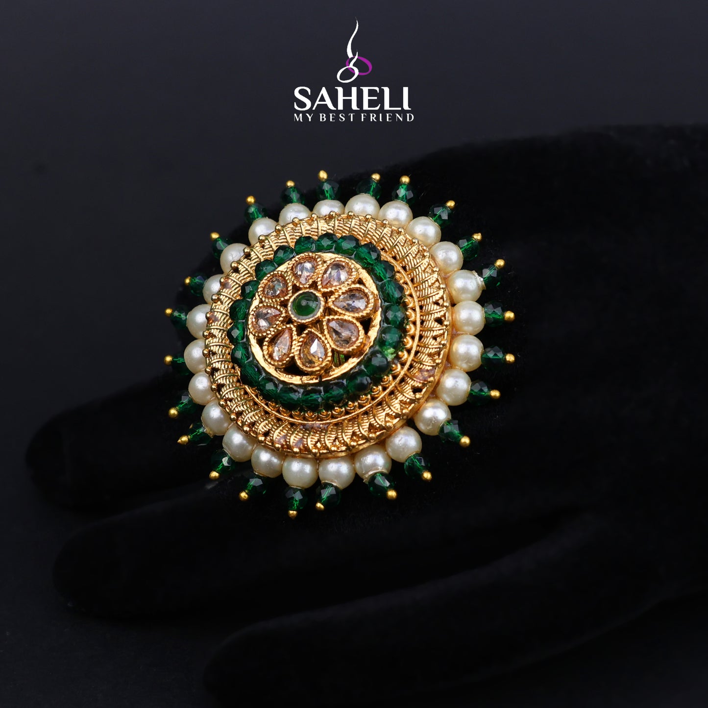 Gold and pearl brooch with green stones on a black background, featuring the brand 'Saheli'.