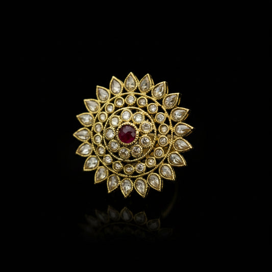 A gold-toned Indian style Kundan choker brooch featuring a central red ruby stone surrounded by clear stones in a sunburst pattern, displayed on a dark gray reflective surface.