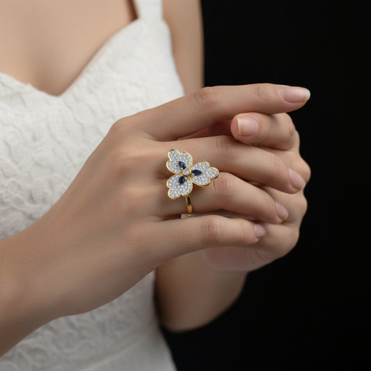 Gold flower-shaped ring with gemstones on a gray background, featuring the brand 'SAHELLI'.