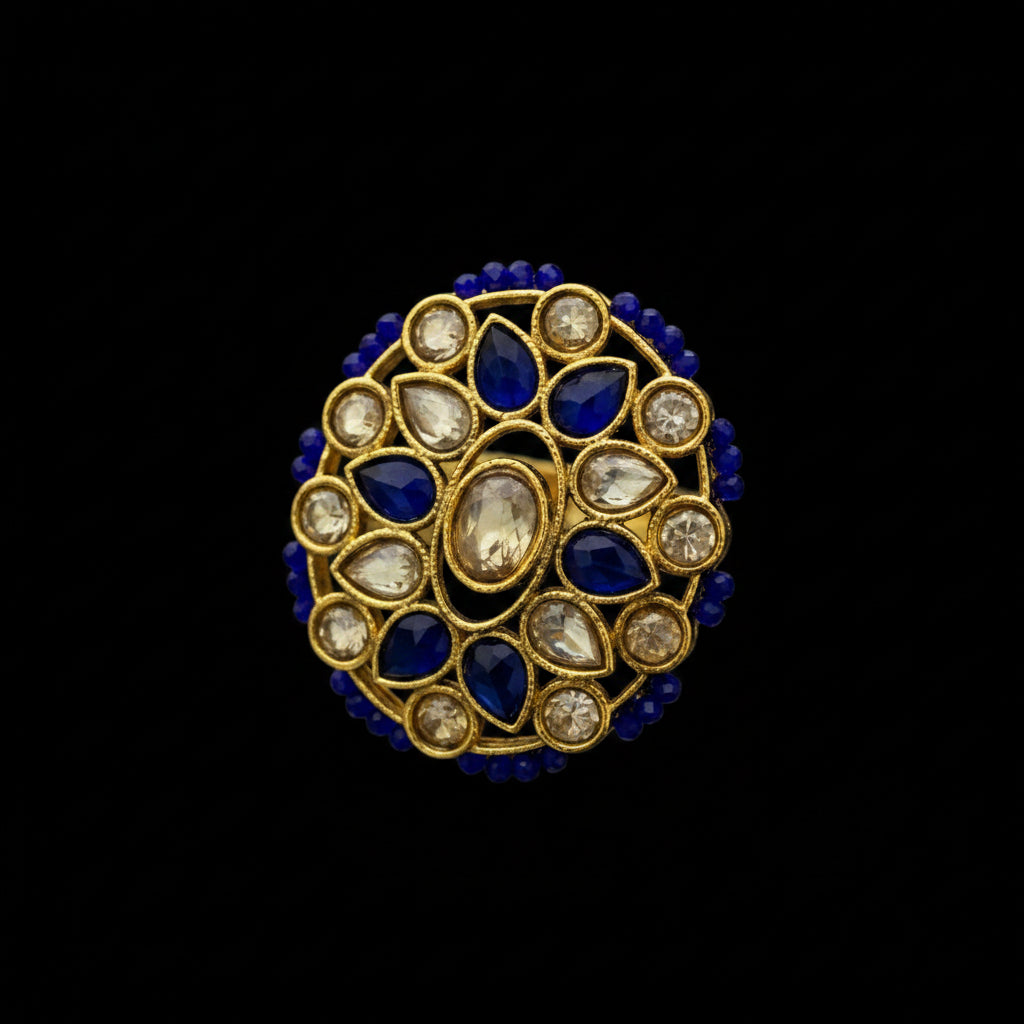 A traditional Indian gold ring featuring a circular floral mandarin design with deep blue stones and clear stone accents, set against a dark gray background.