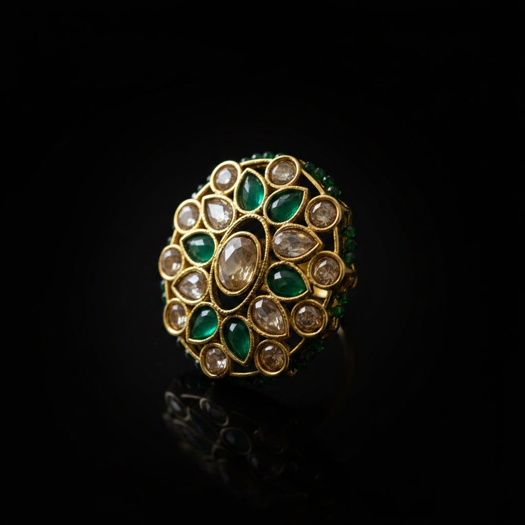 A gold-plated traditional Indian Kundan ring featuring green stones and clear stones, photographed on a dark reflective surface. The SAHEU brand logo is visible in the top left corner.