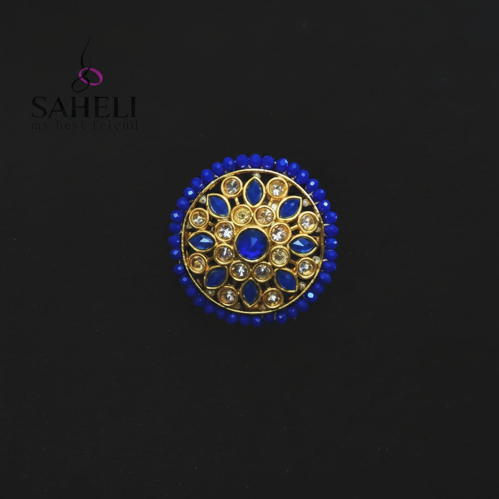 A circular gold-toned brooch or decorative pin featuring a central blue stone and surrounding blue and clear stones, displayed on a dark background with the SAHELI brand logo in the upper left.