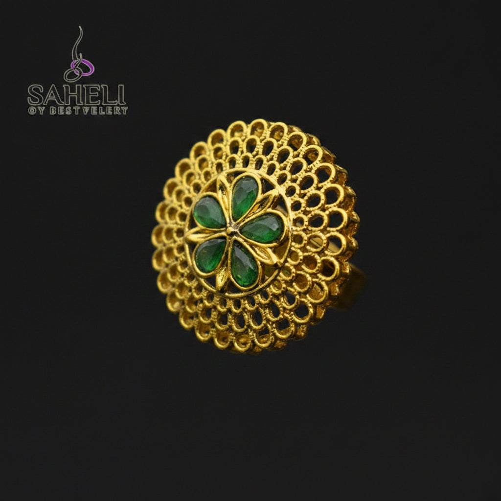 A gold filigree ring with a central four-petal floral motif in green enamel, displayed on a dark reflective surface. The Saheli brand logo is visible in the top-left corner.