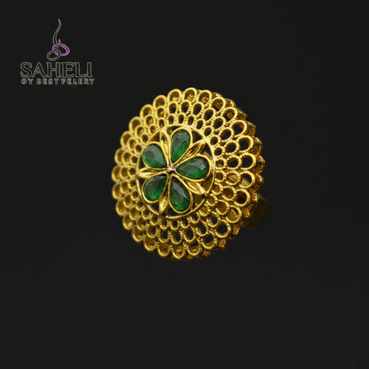 A gold filigree ring with a central four-petal floral motif in green enamel, displayed on a dark reflective surface. The Saheli brand logo is visible in the top-left corner.
