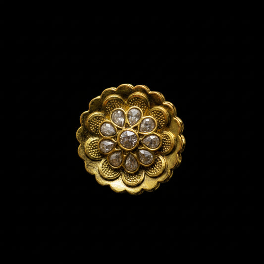A gold ring with a large, ornate lotus-shaped floral face set with small clear stones, photographed on a dark gray reflective surface. The Saheli brand logo is visible in the top-left corner.